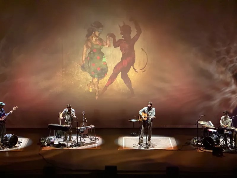 The band performing under warm amber and red stage lighting, with a large projected illustration behind them of a woman in a polka-dot dress dancing hand-in-hand with a red devil.