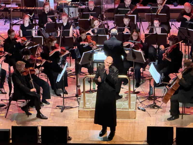 Billy Corgan, bald and dressed in a long black coat, singing into a microphone at the conductor's podium with the orchestra arranged behind him.