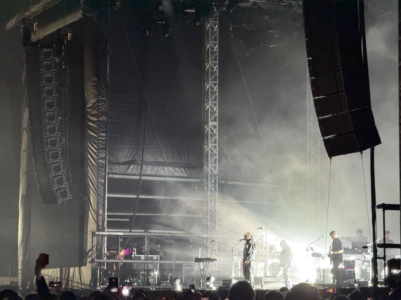 A music performance stage with musicians illuminated by bright lights, surrounded by smoke and a large sound system.