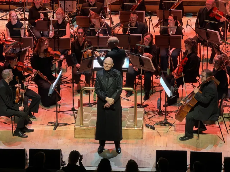 Billy Corgan standing at the podium with hands clasped, looking out toward the audience, orchestra and choir behind him bathed in red and pink light.