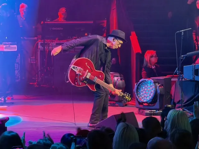 A Garbage guitarist in a hat bending low with a red semi-hollow electric guitar under pink and blue stage lighting, with the drummer and another band member visible in the background.