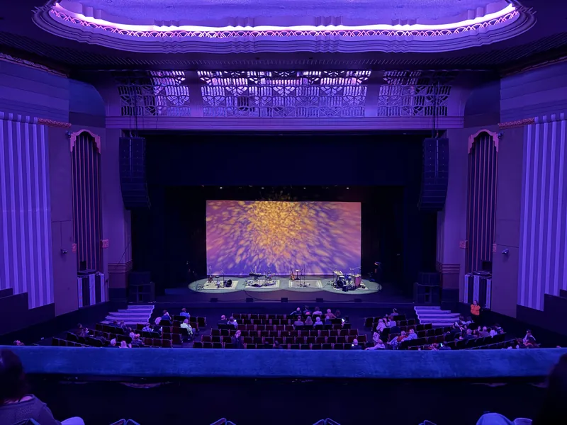 Wide view from the upper balcony of an ornate art-deco theatre before the show, with a warm gold burst of light projected across the backdrop and the empty stage set up with instruments on four spotlit rugs.