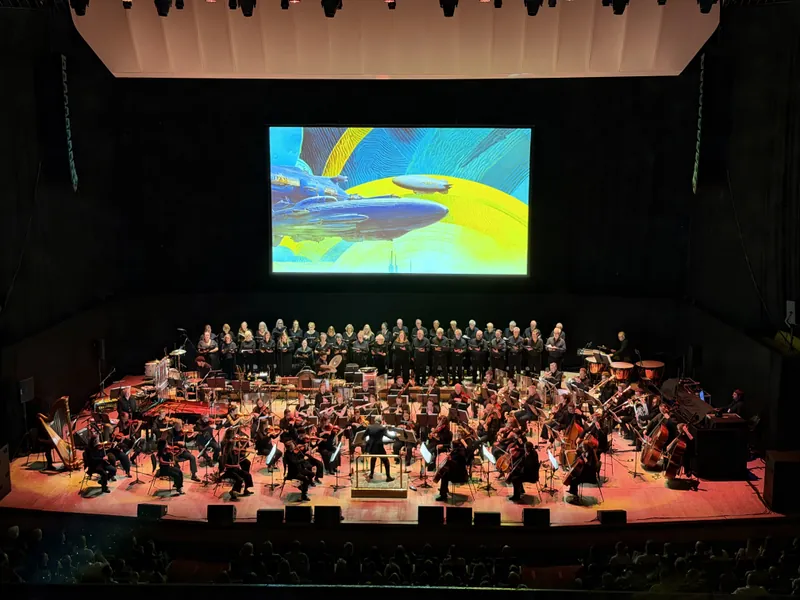 Another wide view of the orchestra and choir on stage, this time under a retro-futuristic illustration of airships drifting past a yellow planet projected on the big screen.