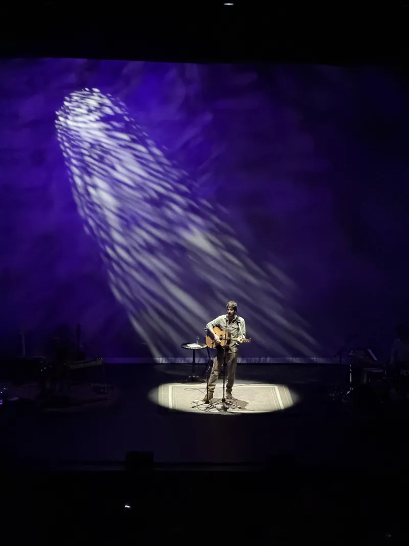 The frontman alone in a spotlight playing acoustic guitar and singing, dramatic white and purple light beams streaking across the deep blue backdrop behind him.
