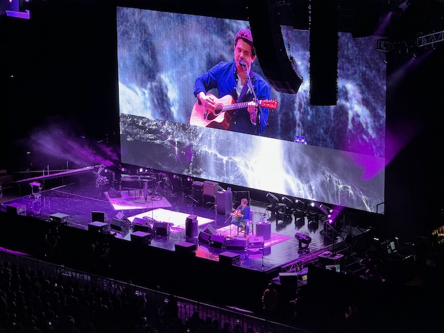 John sitting on a chair stage left playing an acoustic guitar and singing