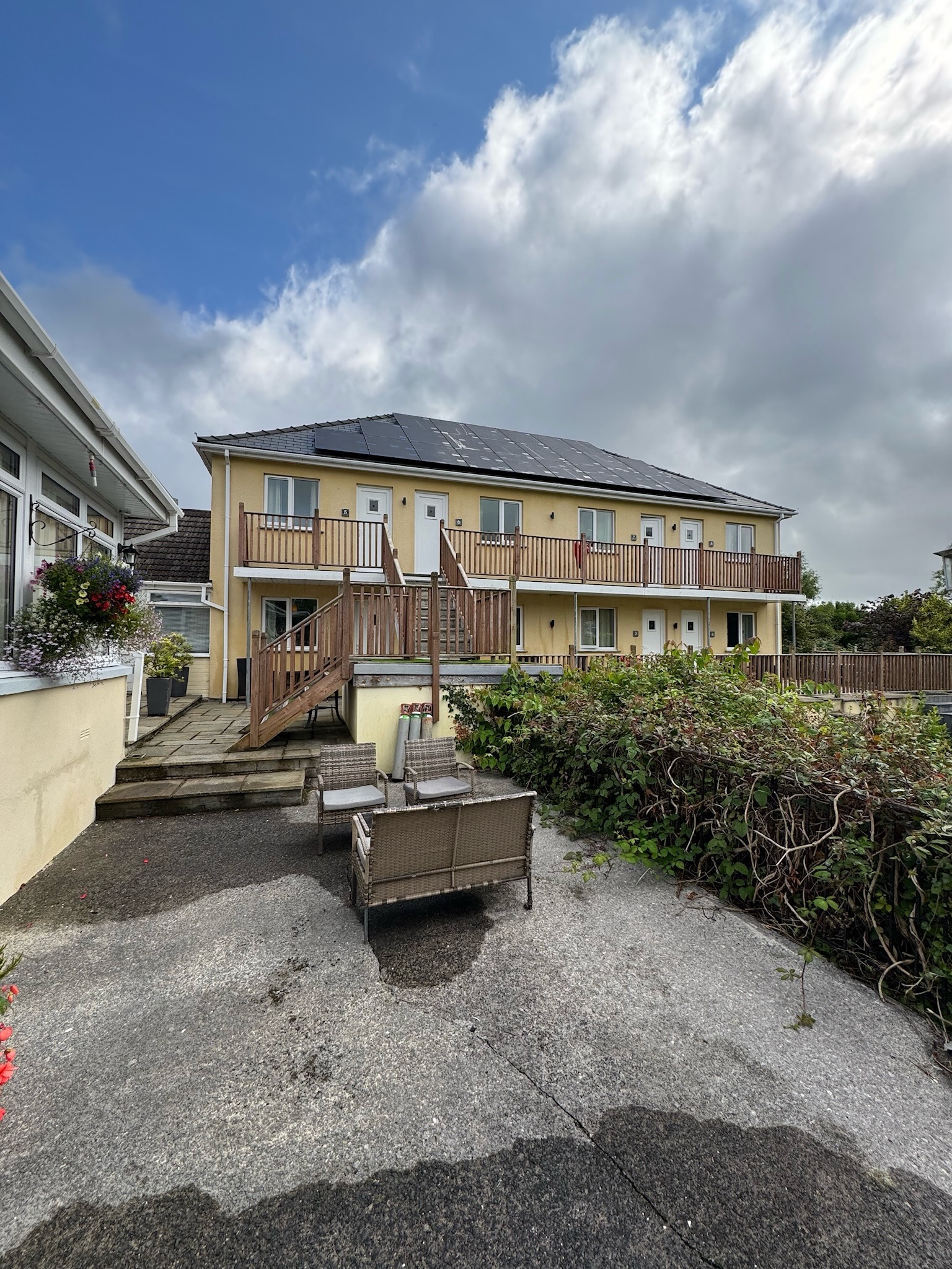 Outside of The Manian. A tan 2 storey building with a lush green garden in front along side a patio.