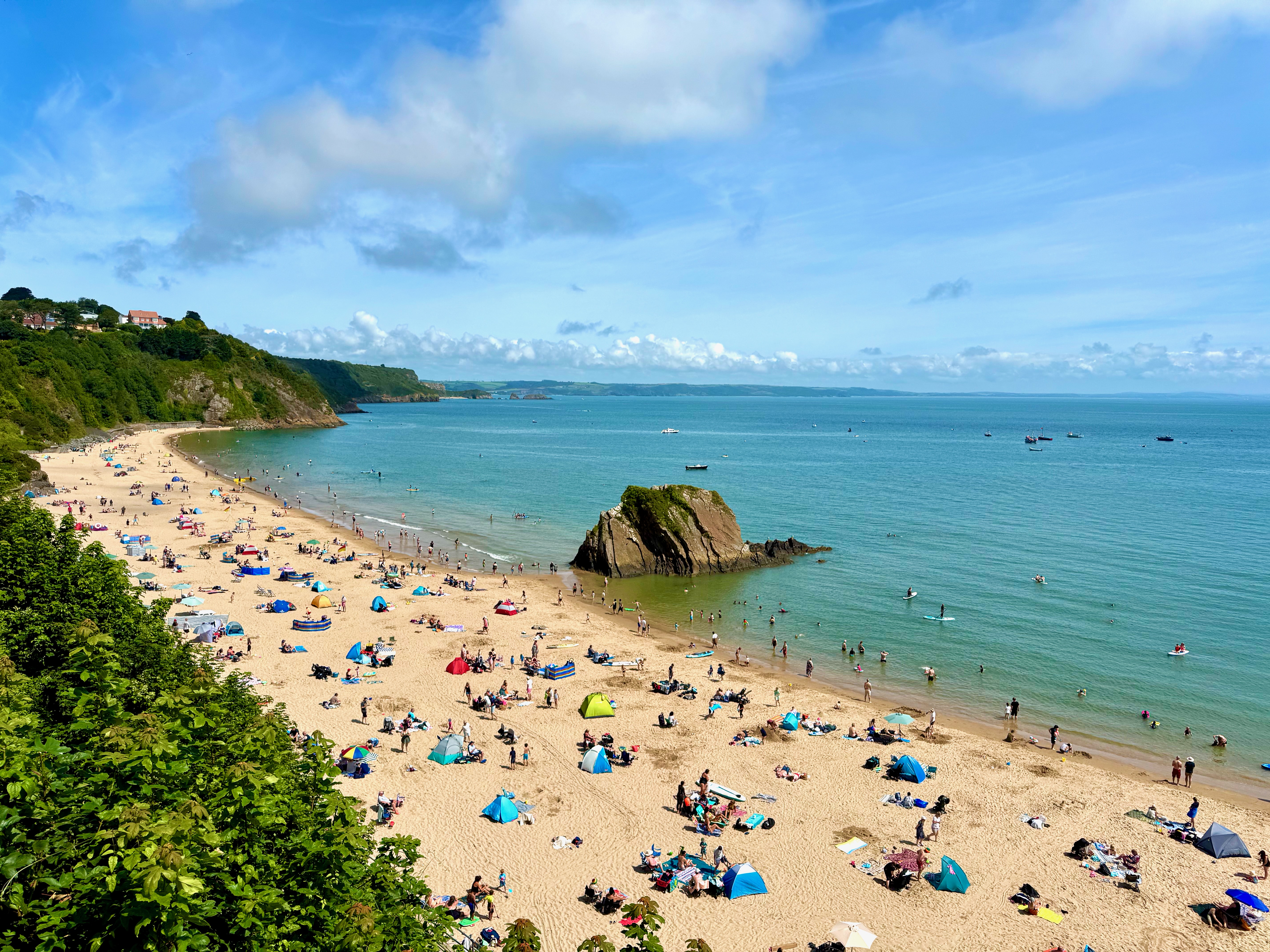 A view down the beach back towards Saundersfoot. The beach is full of people enjoying the sun and playing in the water. Just off the beach in the water a giant rocky crag is sticking out.