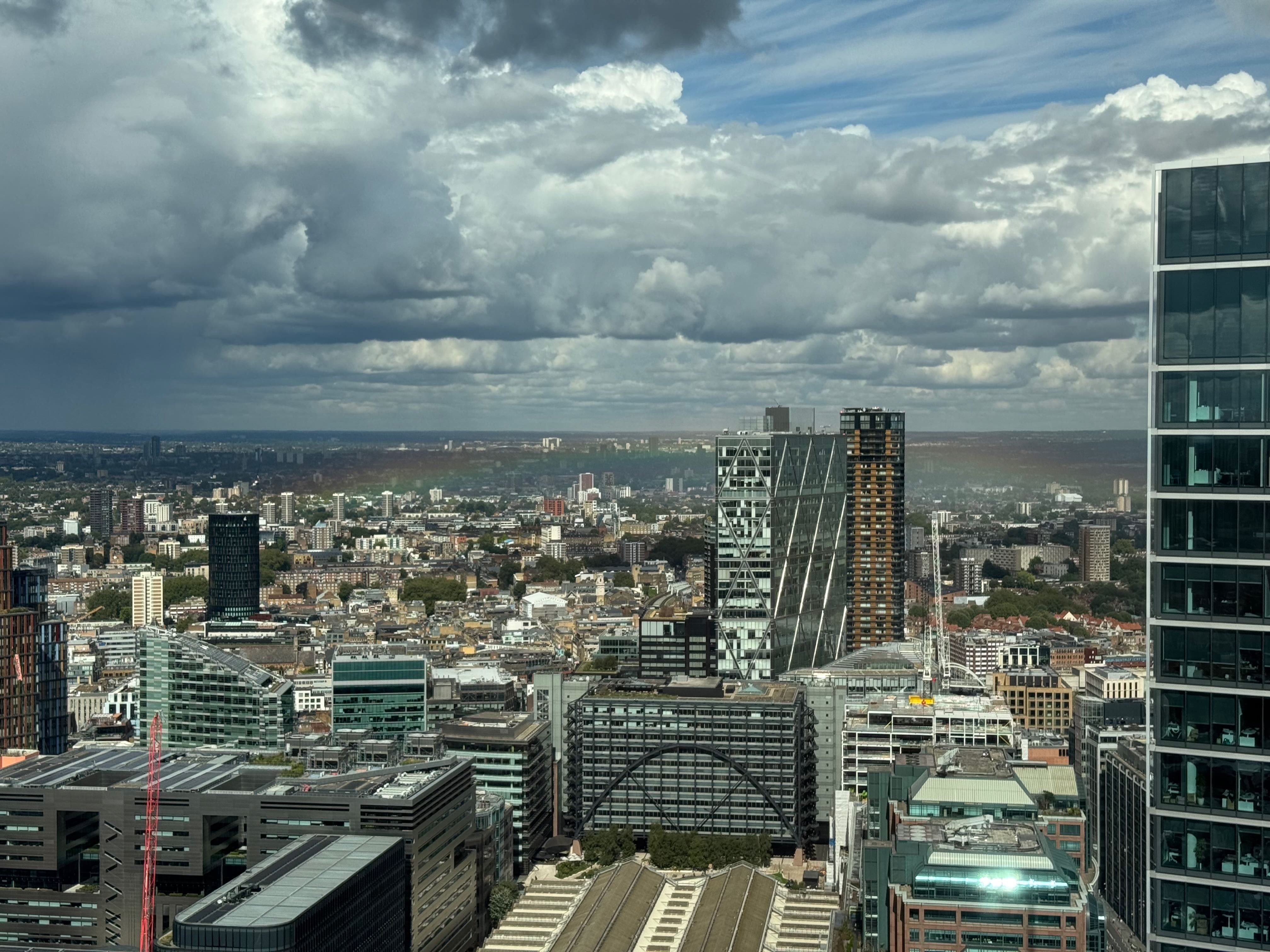 From high up looking over east London with a lot of clouds and a faint rainbow showing around the buildings.