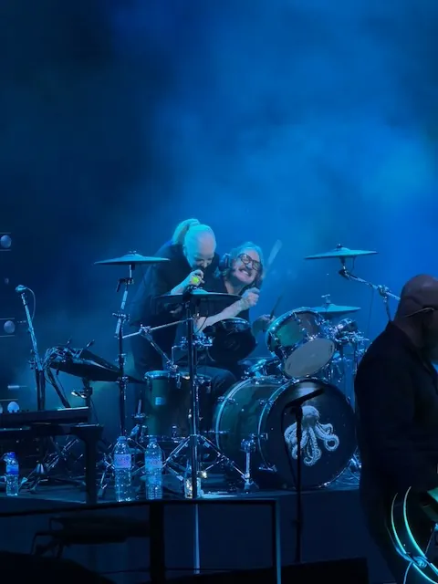 Shirley Manson leaning in towards the drummer, both appearing to share a moment mid-song, illuminated by blue haze. The bass drum has the band's octopus logo on it.