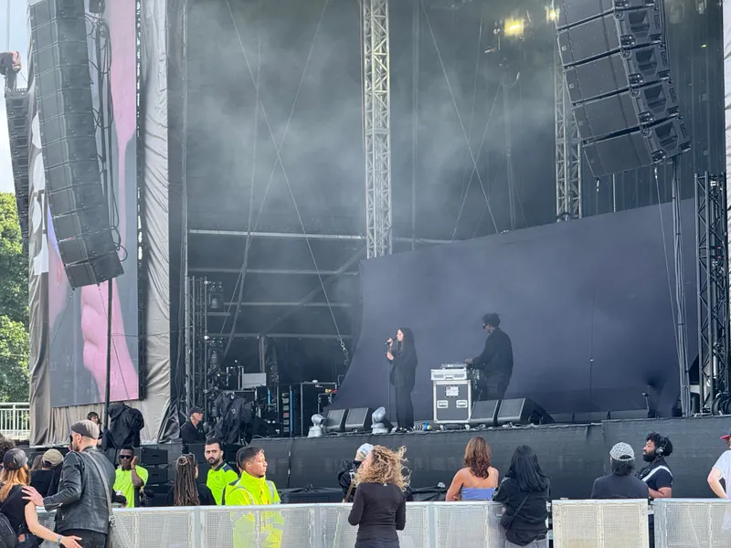 A performer sings on an outdoor concert stage with a DJ beside them, while security staff and audience members stand in front of the stage.