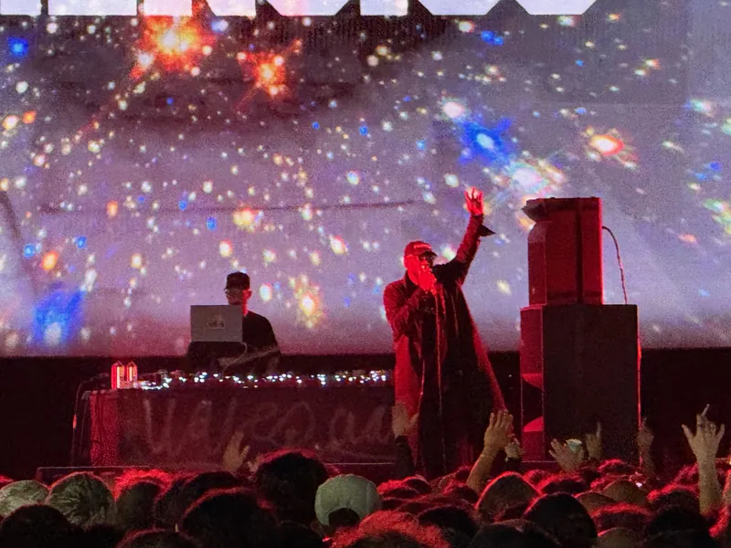 A performer on stage, microphone in hand, with a crowd of people in the foreground and a starry backdrop projected onto a wall.