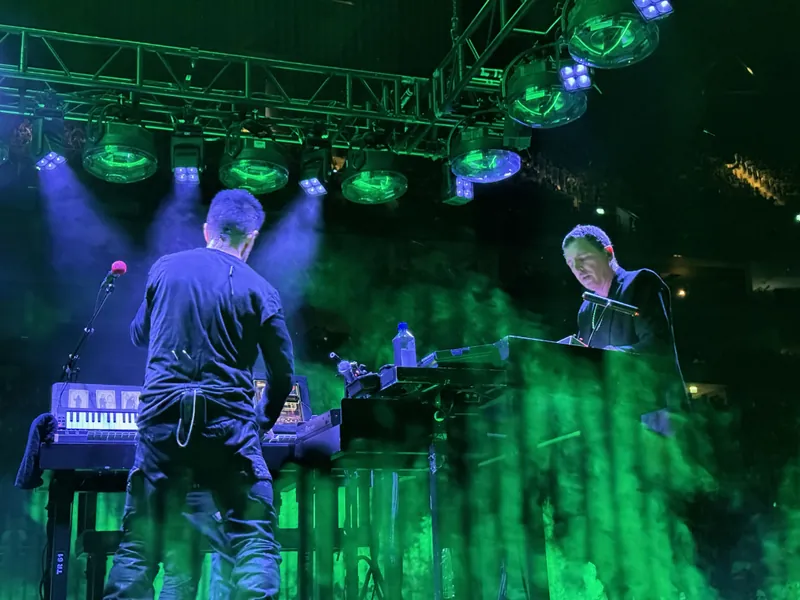 Close up of Trent and Atticus on the second stage. Trent has his back to the camera and you can see Atticus in profile. There are green columns of light coming up from the edge of the stage.