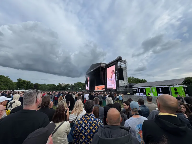 Large crowd at a music festival, watching a performer on a stage with a large screen, under a cloudy sky.