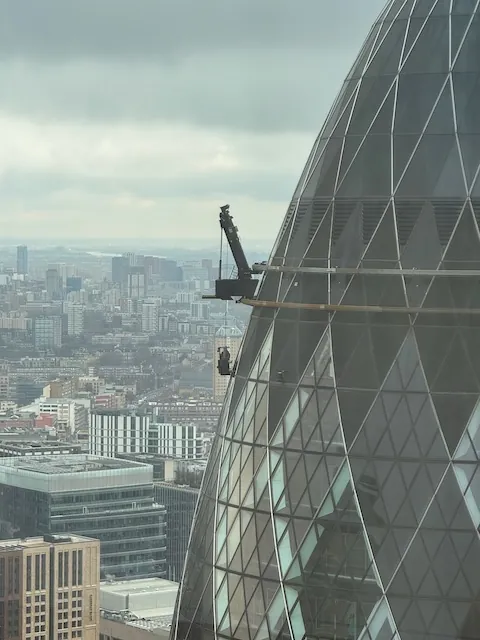A crane attached to the side of the gherkin in London with someone hanging down washing windows