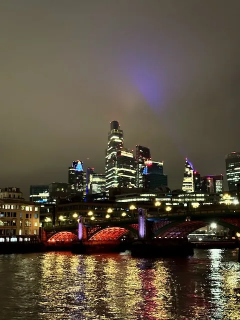 Looking across the Thames to the skyscrapers in the City of London all lit up at night and casting different coloured beams into the low clouds