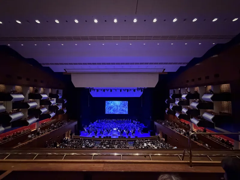 Wide view of the concert hall from the upper tier, with a full orchestra and standing choir on a blue-lit stage beneath a screen showing an abstract blue constellation pattern, as the audience settles in.