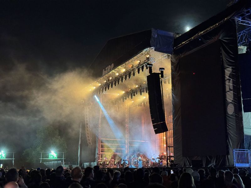 A nighttime concert scene with a large stage illuminated by colorful lights, surrounded by an audience in a misty atmosphere.