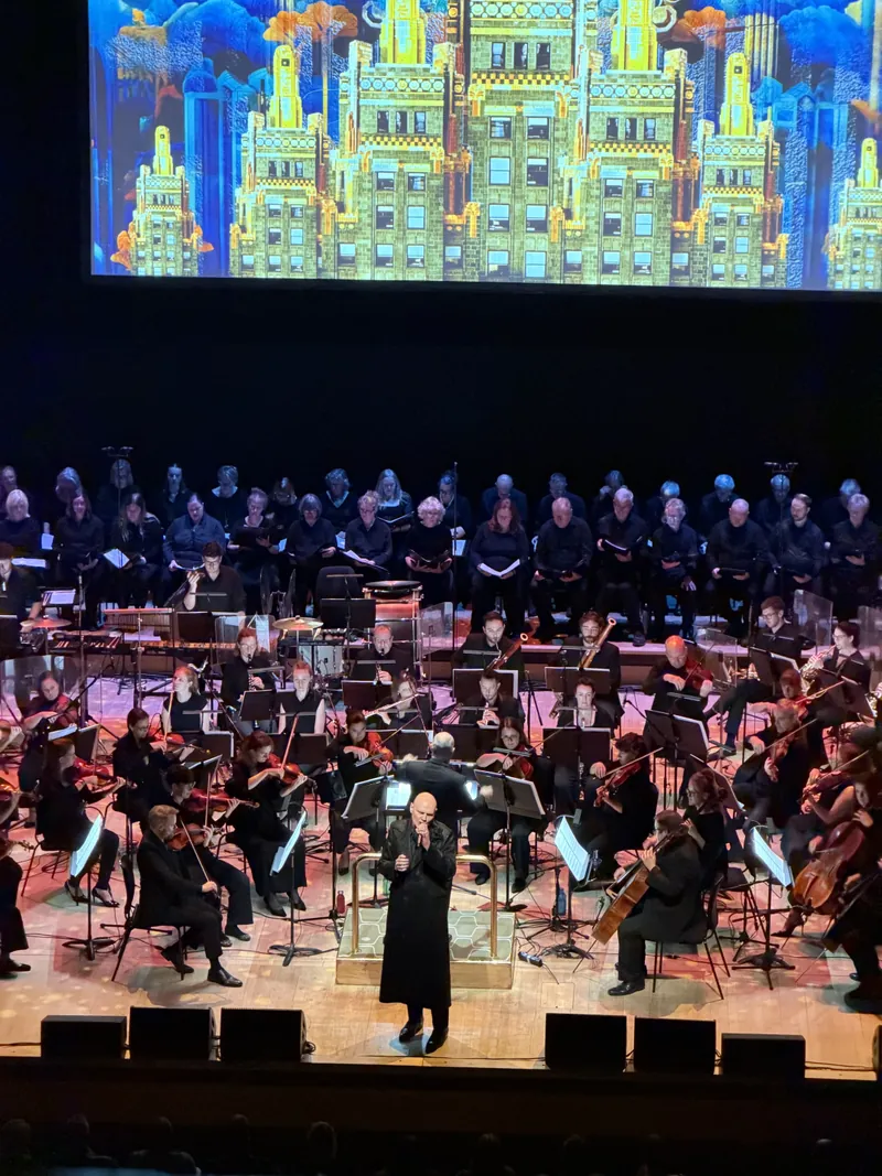 Billy Corgan at the podium mic with the choir standing behind the orchestra, a glowing art-deco cityscape illustration filling the screen above the stage.