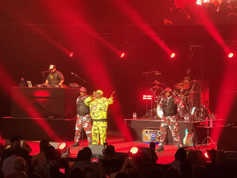 Flava Flav on stage in yellow camo with two other men standing at attention on either side of him in red and white camo wearing bullet proof vests.