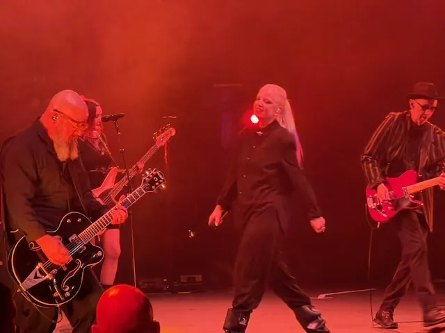 The full band performing under deep red stage lighting β a guitarist with a black Gretsch on the left, Shirley Manson dancing at centre stage, and a guitarist with a red guitar on the right, with a backing vocalist visible at the rear.