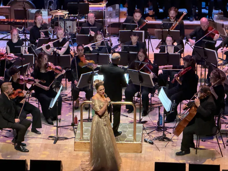 A guest vocalist in a shimmering gold gown singing into a handheld microphone at the conductor's podium, surrounded by the orchestra's string section.
