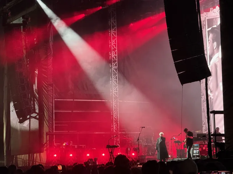 A performer stands on stage under red lights, with a large projected image behind them and musicians playing instruments nearby.