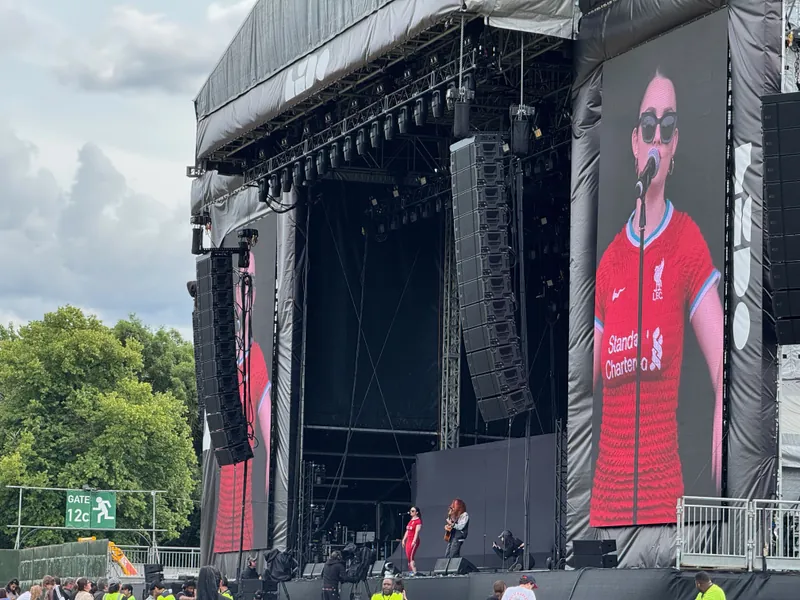 A prominent stage is set with a large screen showing a woman in a red shirt, actively addressing the audience.