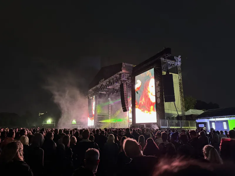 A crowd gathers in front of a large concert stage at night, featuring bright screens and atmospheric smoke effects.