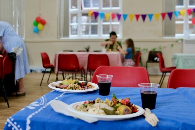 Two plate of food sitting on a table with a blue table cloth in a church hall which is decorated for a party