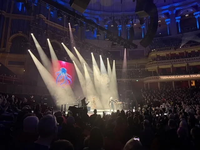 Garbage performing on stage at the Royal Albert Hall, with a glowing blue octopus projected on the LED screen and dramatic white spotlights cutting through haze. A "Teenage Cancer Trust" banner is visible on the balcony.