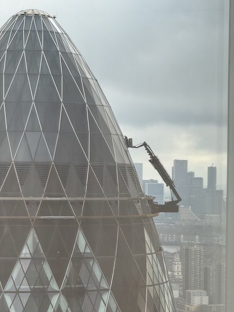 A crane attached to the side of the gherkin in London that is lifting someone up to the top of the building to wash windows