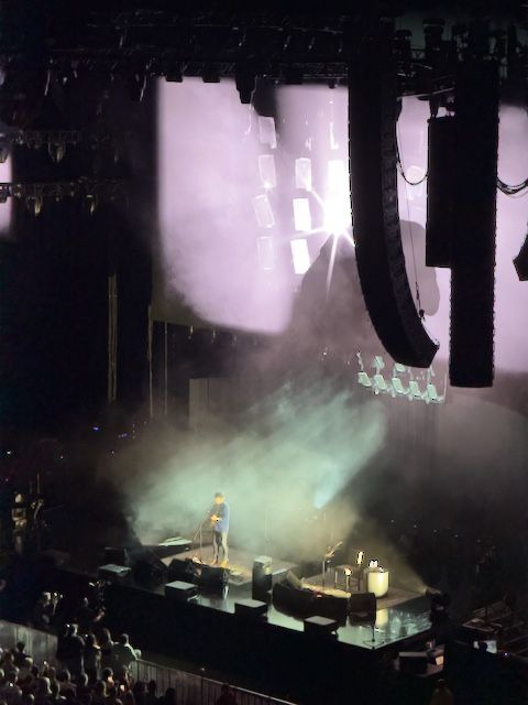 John standing center stage at a microphone holding his acoustic guitar. Bright white lights shining down on him from behind