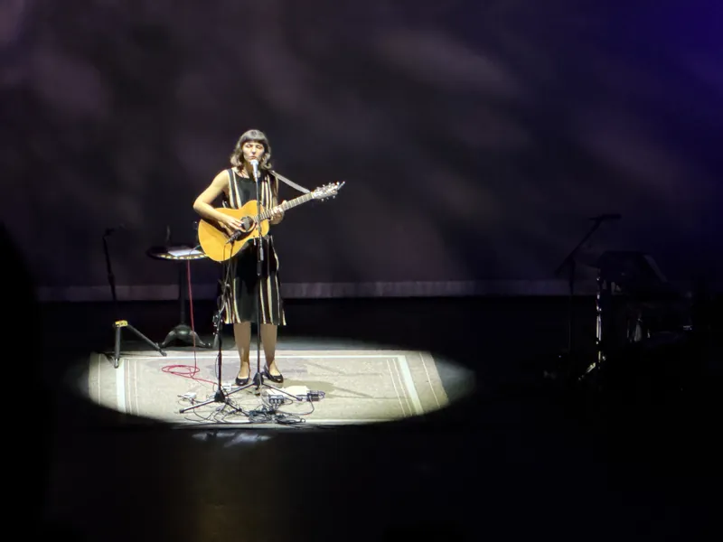 Natalie Jane Hill in a striped dress playing acoustic guitar and singing into a microphone, standing alone in a single spotlight on a patterned rug.