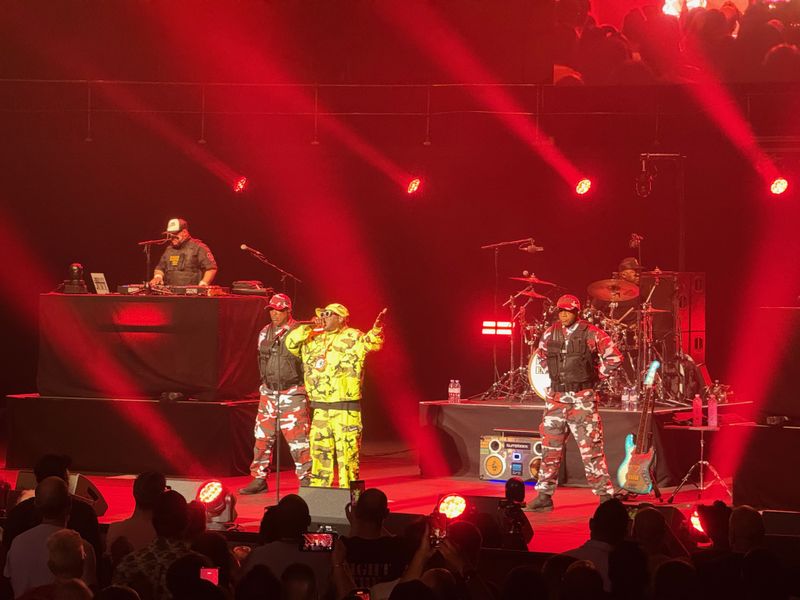 Flava Flav on stage in yellow camo with two other men standing at attention on either side of him in red and white camo wearing bullet proof vests.