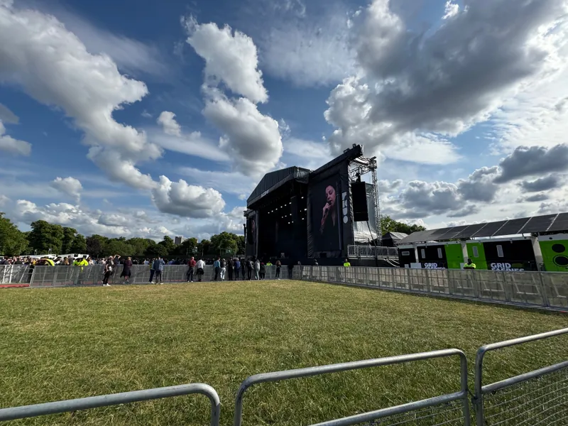 A captivating image displays a bustling concert scene, where a towering black stage with a massive screen commands attention. The crowd, eagerly awaiting performance, is surrounded by a fence, against a serene blue sky. The images energy is palpable.