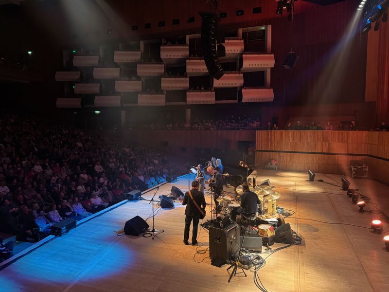 Wider shot of the stage. The young woman from the audience is standing at the microphone singing and the rest of the band is playing.